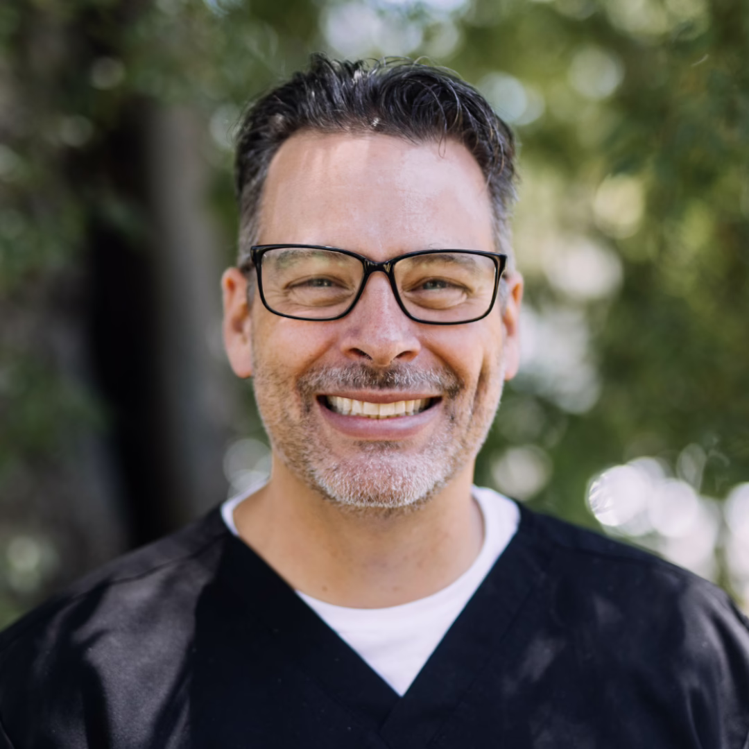 Smiling man in black scrubs and glasses outdoors with greenery in the background, looking at the camera.